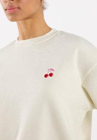 Gros plan sur une personne portant un sweat-shirt beige clair avec un petit design en cerise brodé près de la poitrine et une boucle d'oreille en or.