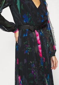 Robe longue noire transparente à manches longues avec imprimé floral bleu, rose et vert vif, jupe plissée et large ceinture noire nouée à la taille.