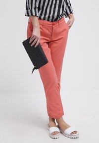 Bawełniane spodnie w kolorze koralowym mają prosty krój, przednie kieszenie i zapięcie na guzik. W połączeniu z płaskimi białymi sandałami i czarnym portfelem.
