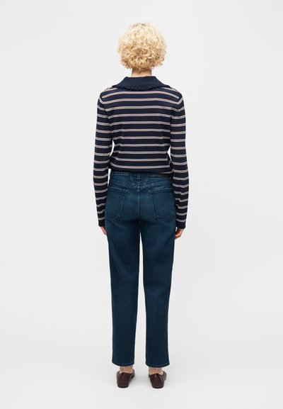 Άτομο με κοντά σγουρά ξανθά μαλλιά, φορόντας μπλε ναυτικό ριγέ μακρυμάνικη μπλούζα, μπλε τζιν και καφέ παπούτσια τύπου slip-on, όρθιο με την πλάτη προς τα εμπρός.