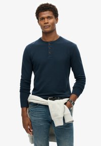 Marineblaues Langarm-Henley-Shirt mit drei braunen Knöpfen, kombiniert mit hellblauen Jeans und einem hellgrauen Sweatshirt, das um die Taille gebunden ist.