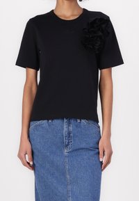 Camiseta negra de manga corta con escote redondo y detalle de volantes texturizados en el hombro izquierdo, combinada con una falda de mezclilla.