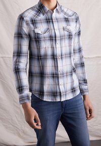 Camicia a maniche lunghe a quadri bianca e nera, con polsini abbottonati e due tasche sul petto. Il tessuto ha una finitura liscia e leggermente strutturata.