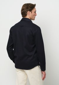Marineblauw wollen shirt met een puntkraag, lange mouwen en zijsplitten. Voorzien van een knoopmanchet en subtiele textuur. Achteraanzicht.