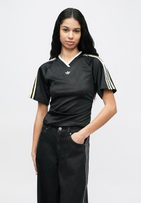 Young woman with long black hair wears a black Adidas shirt with white and yellow stripes and black jeans, posing with one hand in pocket.