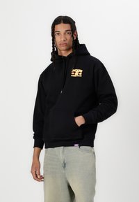 Svart hoodie med frontficka, justerbar dragsko och livlig orange grafisk text "JACKER STREET FOOD." Kombinerad med ljusblå jeans.