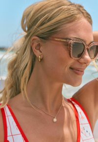 Julge ruudukujuline disain ja täpiline muster prillidel. Näha on blondid juuksed, kuldsed rõngastüüpi kõrvarõngad ja punane mustriga ujumistrikoo.