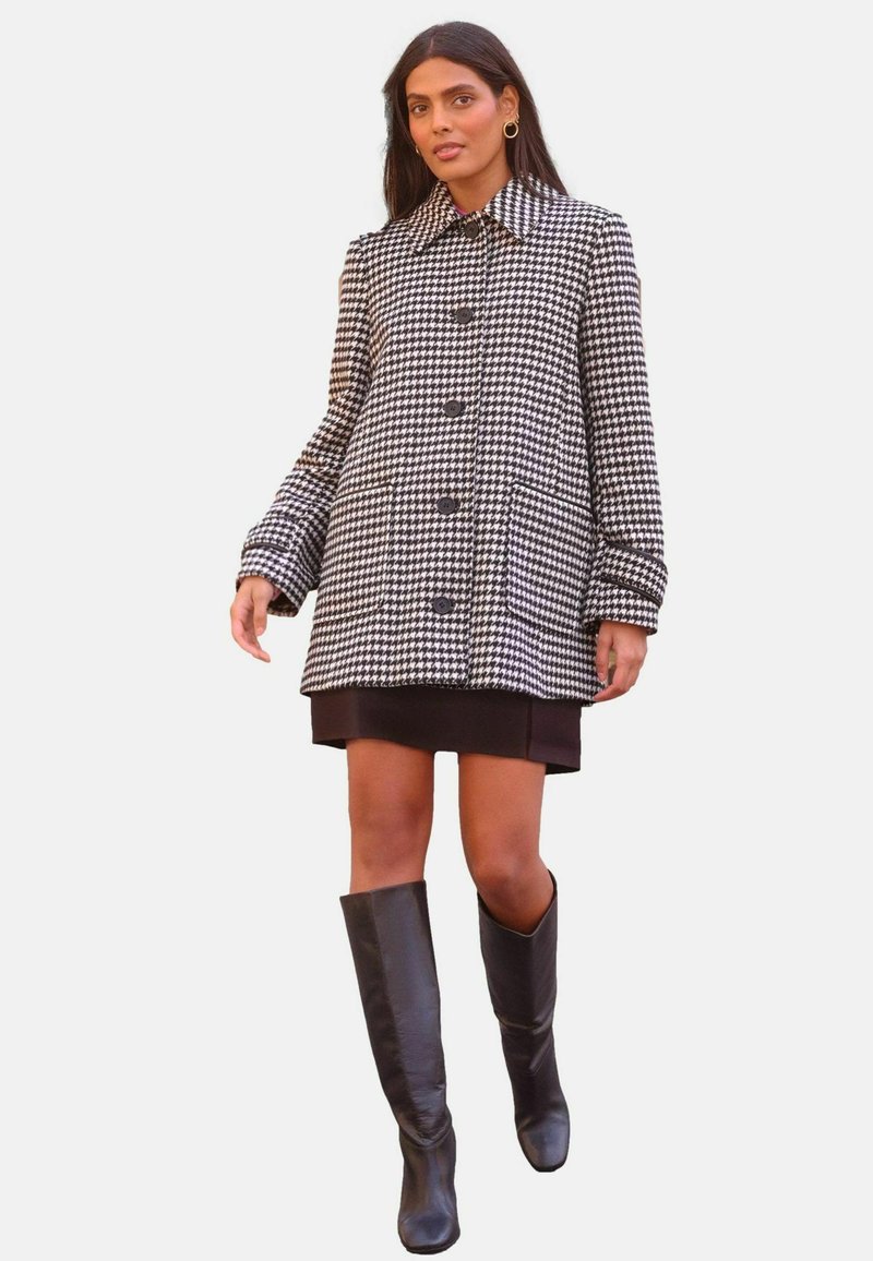 Juoda ir balta houndstooth stiliaus kokybė su apykakle, mygtukų užsegimu ir šoniniais kišenėmis, derinama su trumpa suknele ir juodomis iki kelių aukštakulnėmis bato.