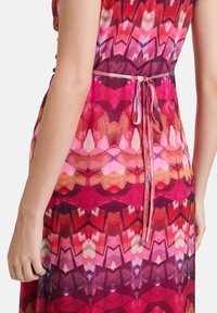 Farbiger Kleid mit einem geometrischen Muster in verschiedenen Schattierungen von Pink, Rot und Violett. Hat ein gebundenes Taillendetail und einen transparenten, fließenden Stoff.