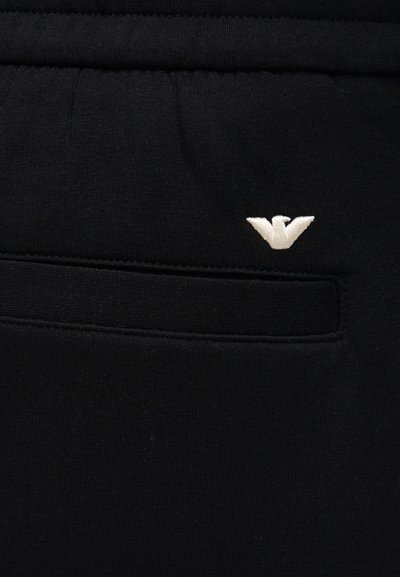 Κοντινό πλάνο μαύρου υφασμάτινου παντελονιού με επίπεδη τσέπη και μικρό λευκό κεντητό λογότυπο αετού πάνω από την τσέπη.