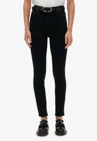 Jeans negros ajustados de tiro alto, combinados con un cinturón negro con hebilla circular, llevados con zapatos oscuros.