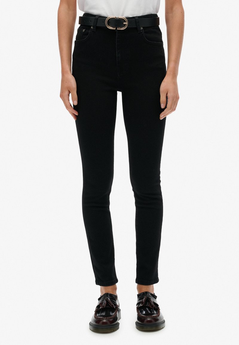 Jeans negros ajustados de tiro alto, combinados con un cinturón negro con hebilla circular, llevados con zapatos oscuros.