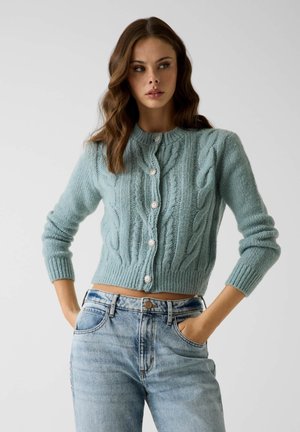 Jeune femme aux cheveux ondulés portant un cardigan bleu clair en maille torsadée avec des boutons en perle et un jean taille haute délavé clair, les mains dans les poches.