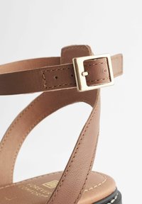 Brun lædersandal med en justerbar ankelrem og en guldfarvet spænde. Sål er sort med synlige syninger.