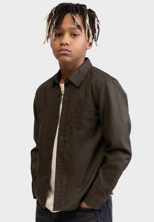 Fiatal személy két színű rasztával, sötét gombos inggel a fehér póló fölött, zsebre tett kézzel álló, egyszerű háttér előtt.