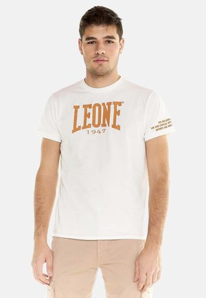 Joven vestido con una camiseta blanca con "LEONE 1947" impresa en marrón, combinado con pantalones cargo beige, de pie contra un fondo liso.