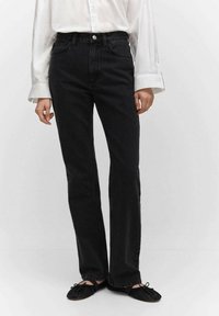 Jeans rectos de mezclilla negra con bolsillos frontales, botón de metal y pasadores para cinturón; combinados con una camisa blanca de manga larga y zapatos planos negros.