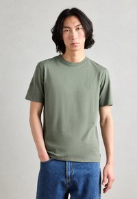 T-shirt en coton vert olive à manches courtes et col rond, décoré d'un petit logo sur la poitrine. Associé à un jean en denim bleu.