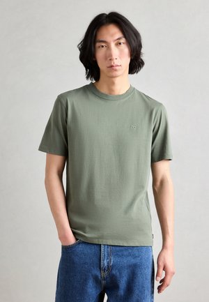 Camiseta de algodón verde oliva con manga corta y escote redondo, que presenta un pequeño logotipo en el pecho. Combinada con jeans de mezclilla azules.