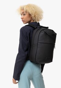 Mochila negra hecha de material suave con un diseño estructurado. Cuenta con correas acolchadas, bolsillo frontal con cremallera y asa de transporte en la parte superior.