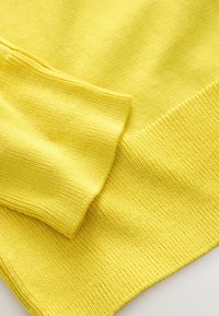 Sárga kötött pulóver bordázott mandzsettákkal és sima textúrával. A szövet könnyű, egyenletes szemkiosztással.