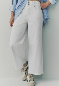 Witte wijde jeans van denim met een hoge taille, voorzien van twee voorkanten zakken en dubbele knoopsluiting. Draagt met beige sneakers.