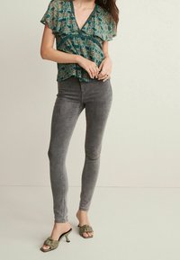 Ärmelloses türkisfarbenes Oberteil mit einem gemusterten Design und Rüschenschichten, kombiniert mit hochtaillierten grauen Skinny-Jeans und olivgrünen Knoten-Mules.