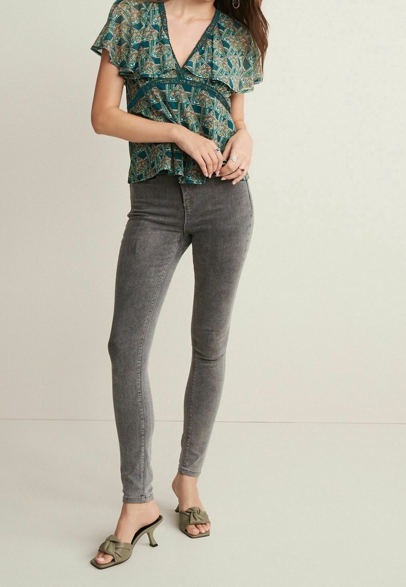 Ärmelloses türkisfarbenes Oberteil mit einem gemusterten Design und Rüschenschichten, kombiniert mit hochtaillierten grauen Skinny-Jeans und olivgrünen Knoten-Mules.