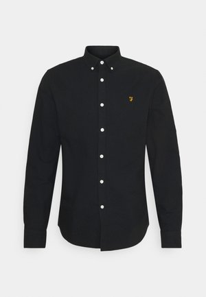 Zwarte blouse met lange mouwen van stof, met een button-down kraag en witte knopen; heeft een klein geel logo op de linkerborst.