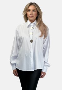 Biała koszula z długimi rękawami, z wyłogowym kołnierzem i dużymi, okrągłymi srebrnymi guzikami z przodu. Gładka tekstura.