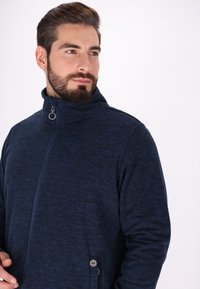 Giacca a zip di colore blu navy con colletto alto, texture morbida e dettaglio dei bottoni laterali. Presenta un sottile motivo melange in tutto il tessuto.