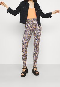Legginsy w kwiaty na fioletowym tle z małymi, wielokolorowymi kwiatami, zestawione z czarną dżinsową kurtką i pomarańczową bluzką. Czarne, ciężkie sandały.