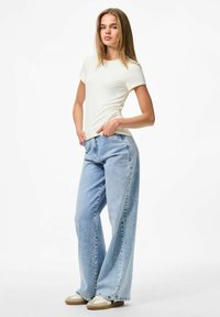 Balta īsām piedurknēm t-krekls savienots ar gaiši ziliem platām kājām džinsiem. Apavi ir bežīgi ar baltām akcentiem. Modelis pozē ar rokām uz gurniem.