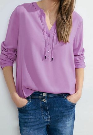 Vrouw met een lavendelkleurige blouse met lange mouwen en een blauwe spijkerbroek, handen in de zakken, staand tegen een effen lichte achtergrond.