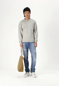 Camisola cinzenta de malha, jeans rasgados azul claro, ténis brancos e uma bolsa tote castanha. Modelo está em pé contra um fundo branco simples.