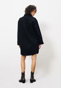 Schwarzes Hemdkleid mit Kragen, langen Ärmeln und seitlichem Schlitz. Kombiniert mit knöchelhohen, schwarzen Stiefeln mit Absatz. Glatte Stoffoberfläche.