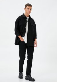 Camisa negra de gran tamaño con dos bolsillos en el pecho, llevada sobre una camiseta blanca, combinada con pantalones negros ajustados y zapatos negros con cordones.