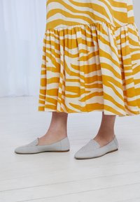 Scarpe slip-on grigie con un design intrecciato sulla parte superiore, abbinate a un vestito a strati giallo e bianco a righe, su un pavimento in legno chiaro.