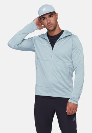Mann trägt hellblaue Zip-up-Jacke und passende Mütze, mit schwarzem Hemd und schwarzer Hose, posiert mit einer Hand hinter dem Kopf.