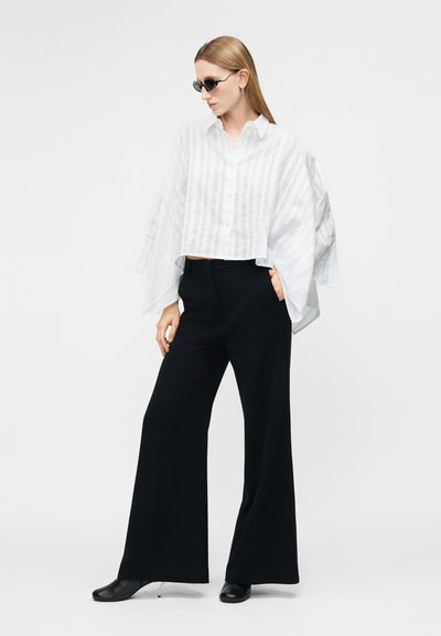 Žena v čiernych širokých nohaviciach, bielej voľnej košeli s dlhým rukávom, čiernych lodičkách na opätku, čiernych slnečných okuliaroch, stojaca s jednou rukou v vrecku.