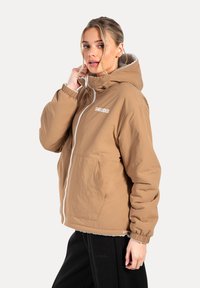 Chaqueta capucha marrón claro con un zipper blanco, puños elásticos y un forro interior suave. Presenta un logo en el pecho y dos bolsillos laterales.