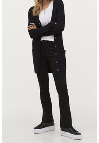 Kvinna står med armarna i kors, iklädd svart knäppt cardigan, vit topp, svarta byxor och svarta plattformsskor mot en enkel bakgrund.