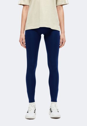 Personne debout portant des leggings bleu marine, des chaussures de sport blanches et un t-shirt beige à manches courtes sur un fond uni.