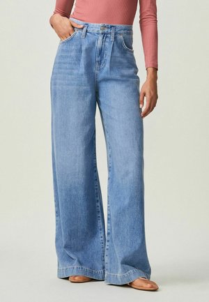 Donna che indossa jeans a vita alta, gamba larga, azzurri chiari e una maglia rosa a maniche lunghe, in piedi con una mano in tasca, scarpe color cuoio visibili.