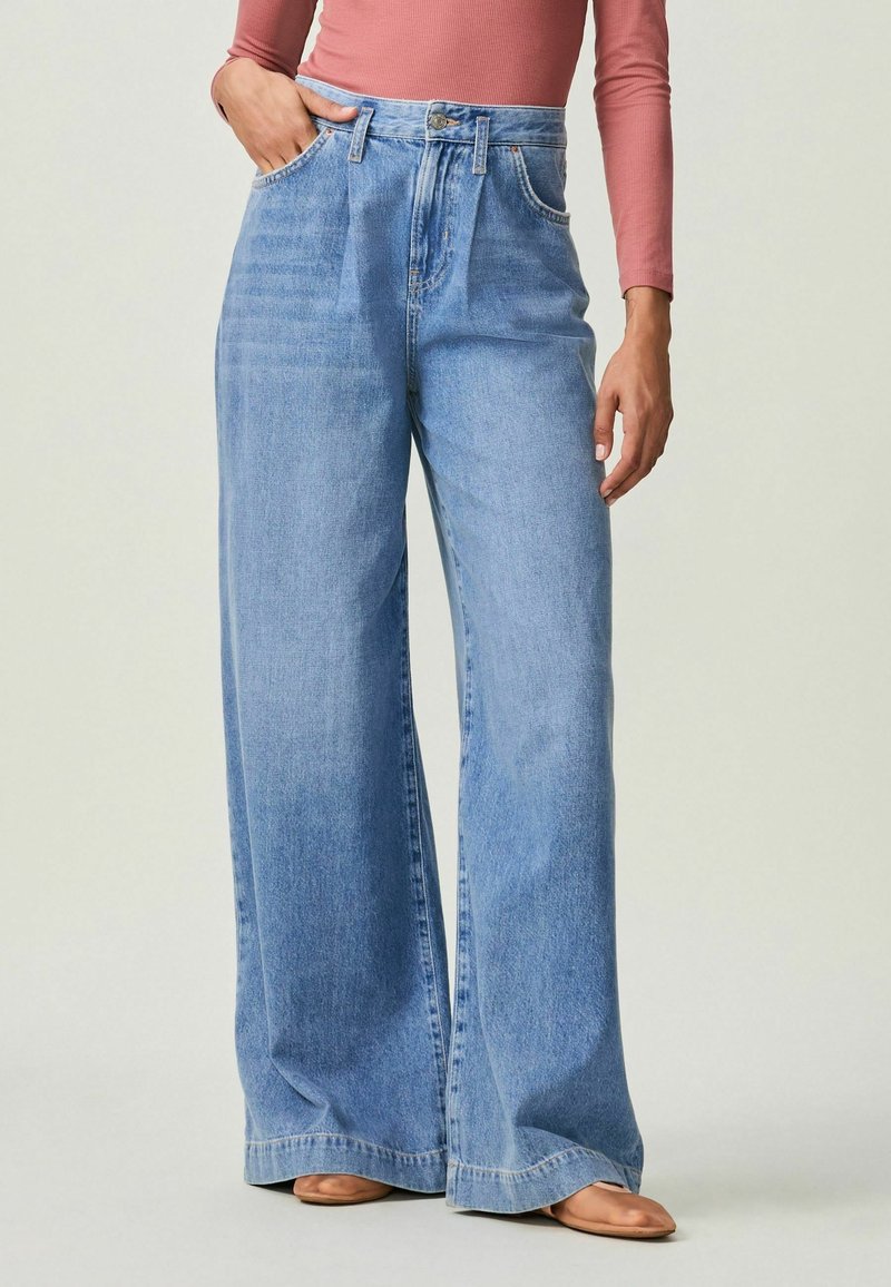 Femme portant un jean taille haute bleu clair et jambes larges, ainsi qu'un haut à manches longues couleur rose, debout avec une main dans la poche, chaussures marron visibles.