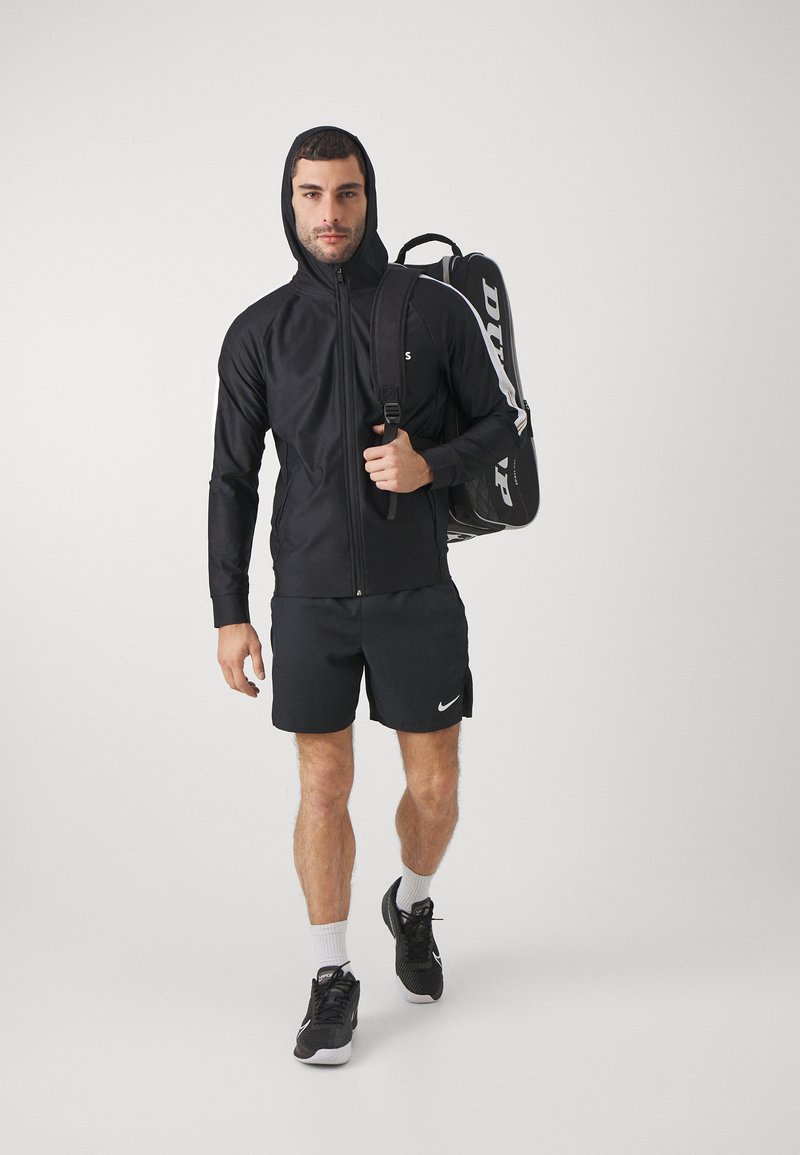 Schwarze Zip-Jacke mit Kapuze, kombiniert mit schwarzen Shorts. Verfügt über ein Logo auf der Brust und einen Rucksack mit Branding an der Seite.