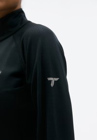 Osoba u crnoj sportskoj majici s dugim rukavima s malim bijelim logotipom na gornjem dijelu ruke na jednostavnoj bijeloj pozadini.