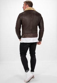 Giacca di pelle marrone con colletto in pelliccia, dettagli trapuntati sulle spalle e sull'orlo, abbinata a jeans neri e sneakers bianche alte.