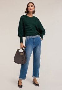 Maglione a coste verde, jeans azzurri chiari con cintura chiodata, décolleté in vernice nera e borsa marrone scuro con dettagli dorati.