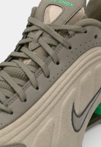 Nike Shox en vert olive atténué, avec une tige en synthétique lisse, des lacets texturés et une semelle rembourrée avec un accent vert visible.